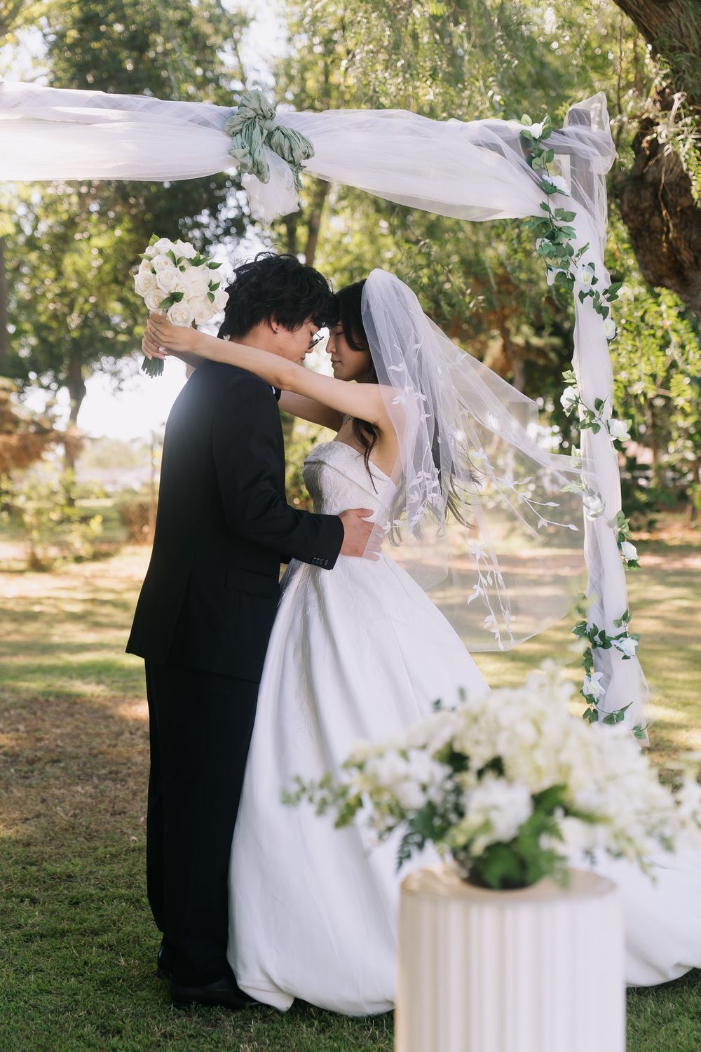 A bride and groom embrace under a floral arch during an outdoor wedding ceremony on a lush green lawn.