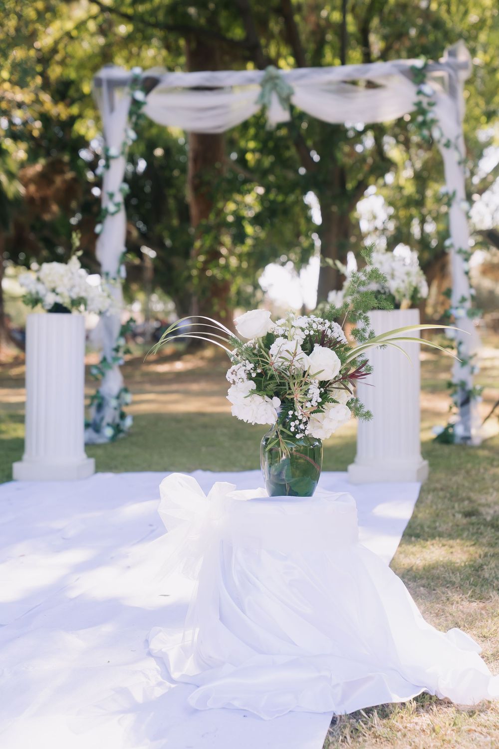 Elegant outdoor wedding ceremony setup with white arch, floral decorations, and bouquet on draped table.
