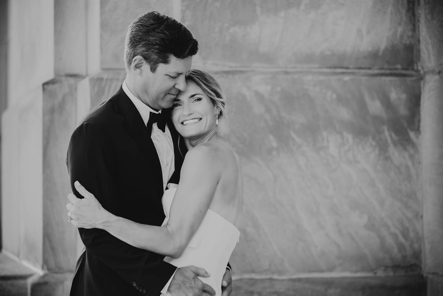 Black and white photo of a couple embracing and smiling on their wedding day near stone columns.