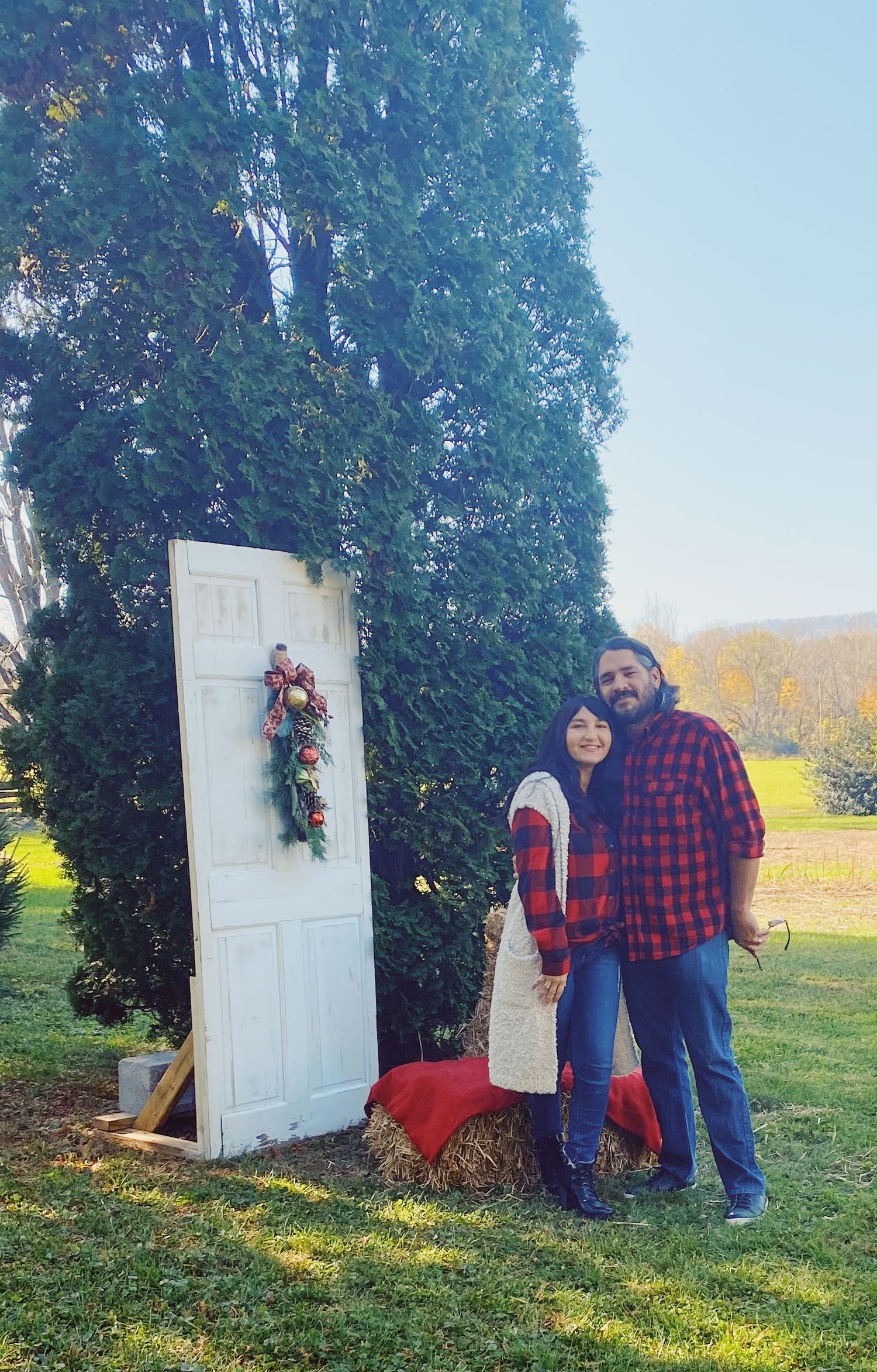 Couple smiling together outdoors beside a white door decorated with a Christmas wreath near a tall evergreen tree.