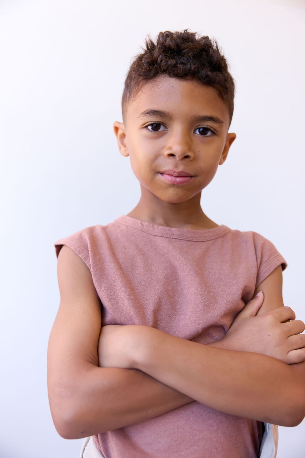 Confident mixed-race boy with arms crossed wearing pink sleeveless shirt against white background.