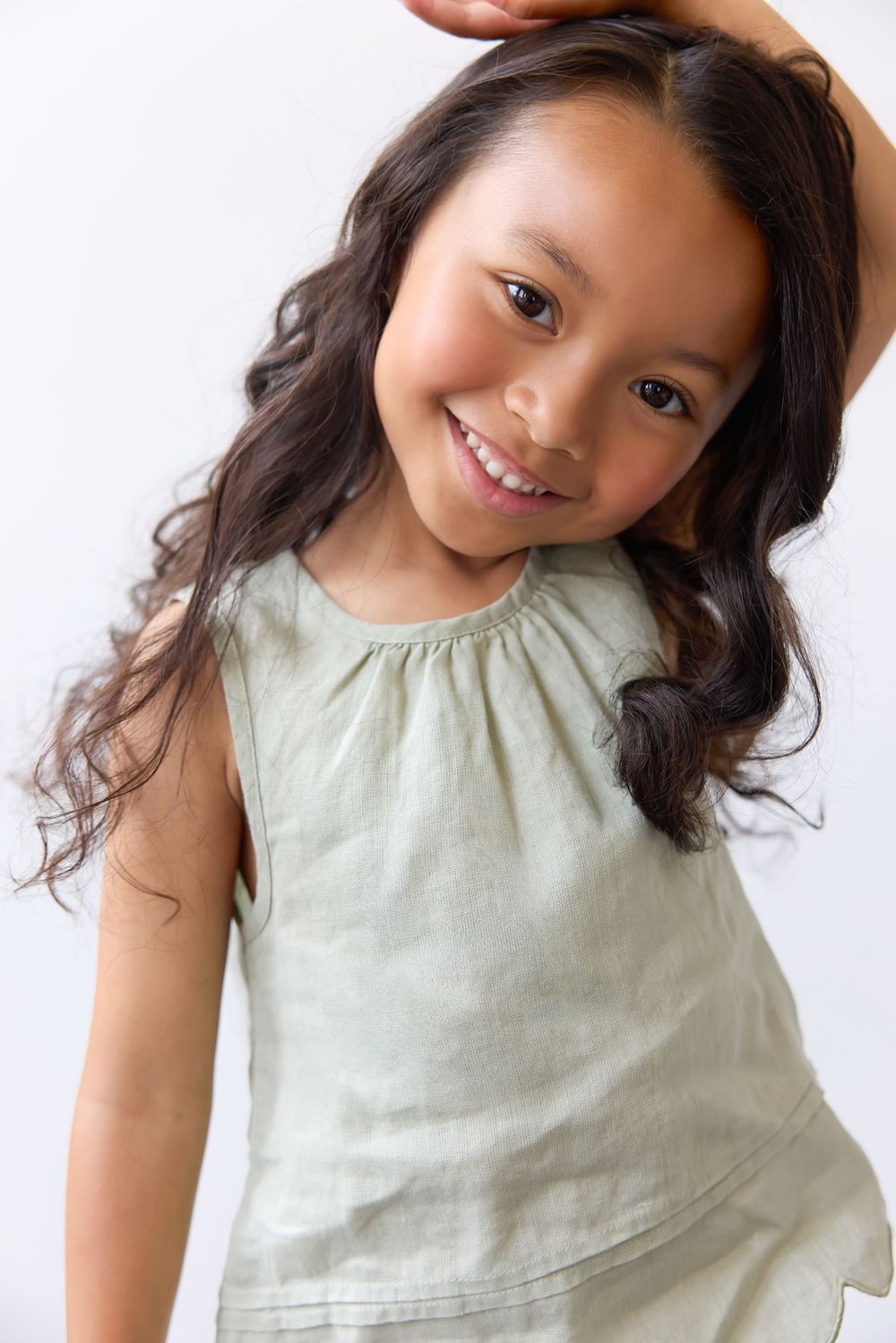 Smiling young Asian girl with long curly hair wearing a sage green sleeveless dress on white background.