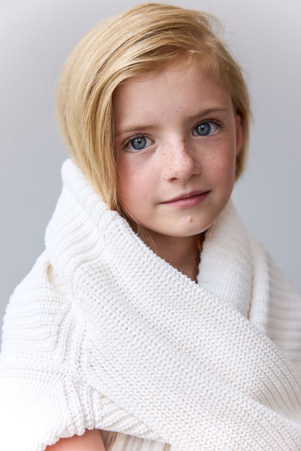 Young blonde girl with blue eyes and freckles wrapped in a white knit blanket.