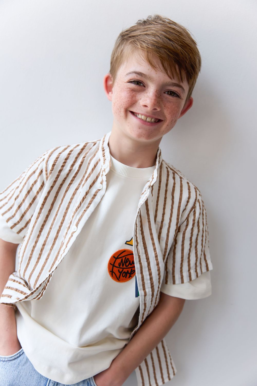 Smiling young boy in striped shirt and graphic tee poses confidently against white background.