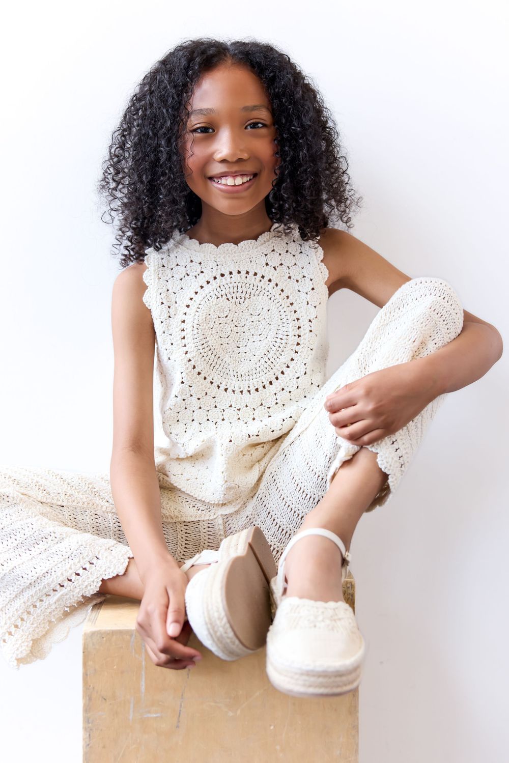 Smiling Black girl with curly hair sitting on box wearing white crochet outfit and espadrille shoes.