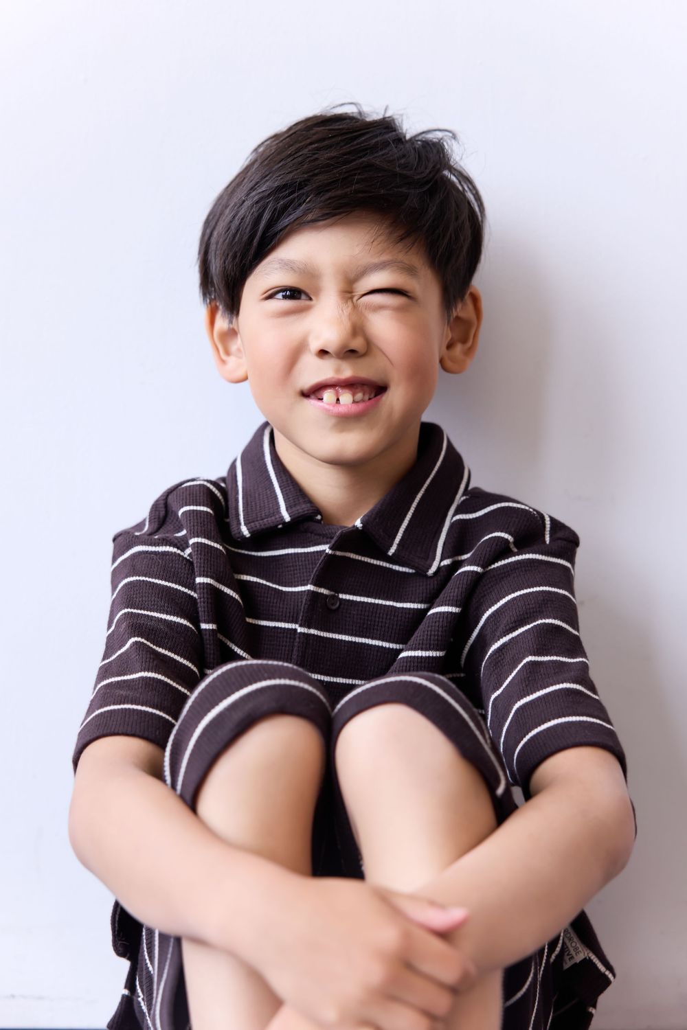 Smiling Asian boy in striped polo shirt sitting with knees up against white wall.
