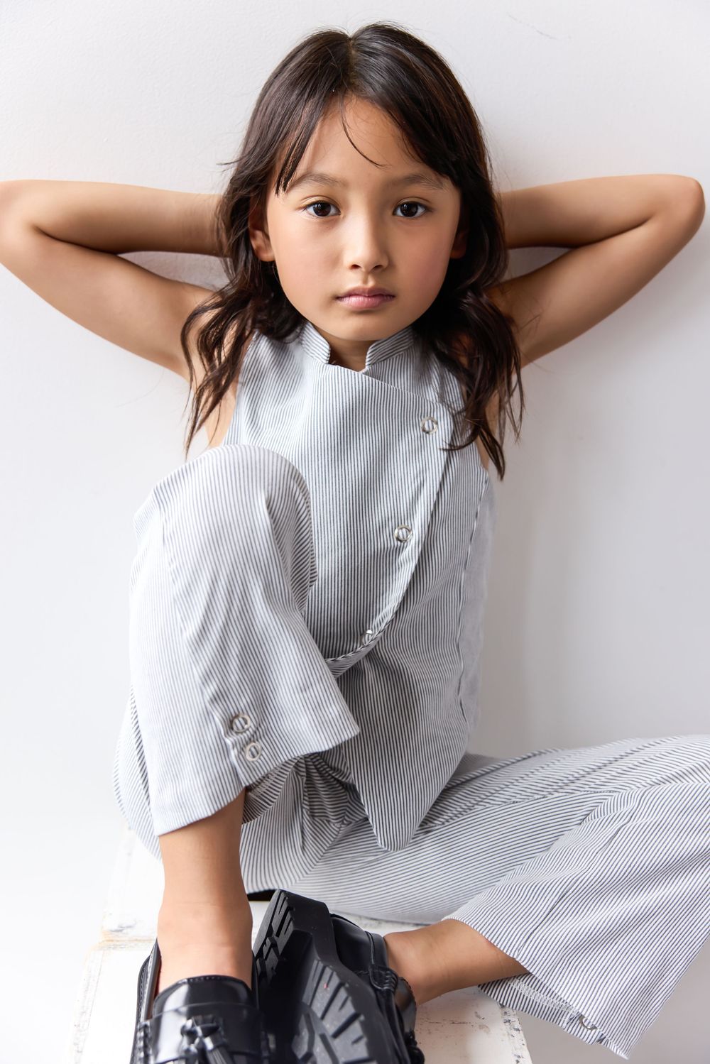 Young girl in striped sleeveless top and wide-leg pants with black shoes, posing against white wall.