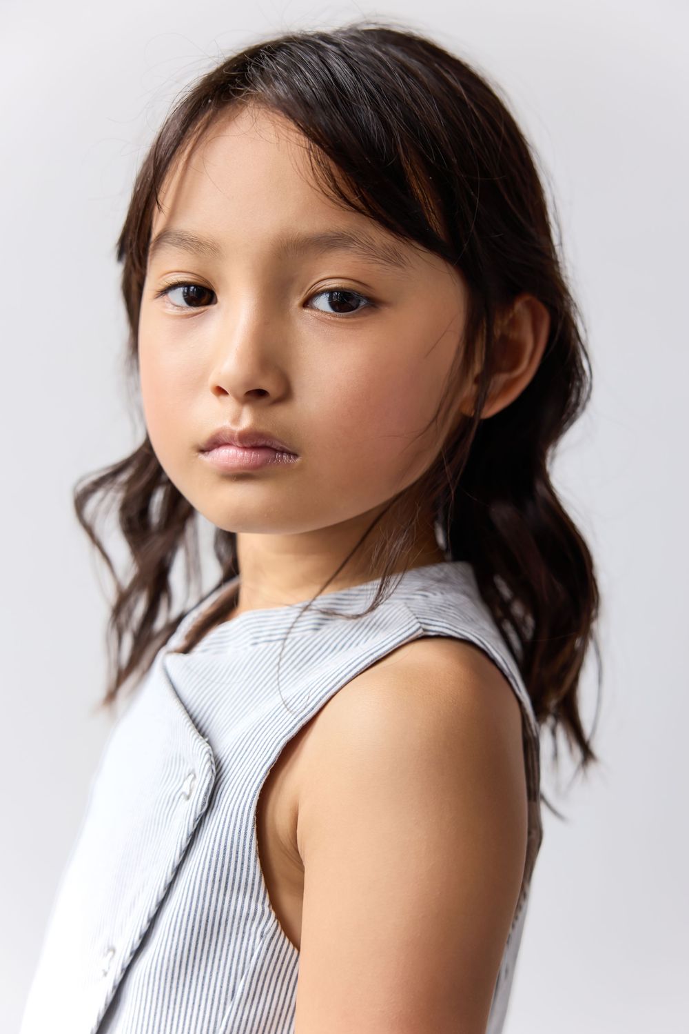 Young girl with dark hair in a striped sleeveless top posing against a light gray background.