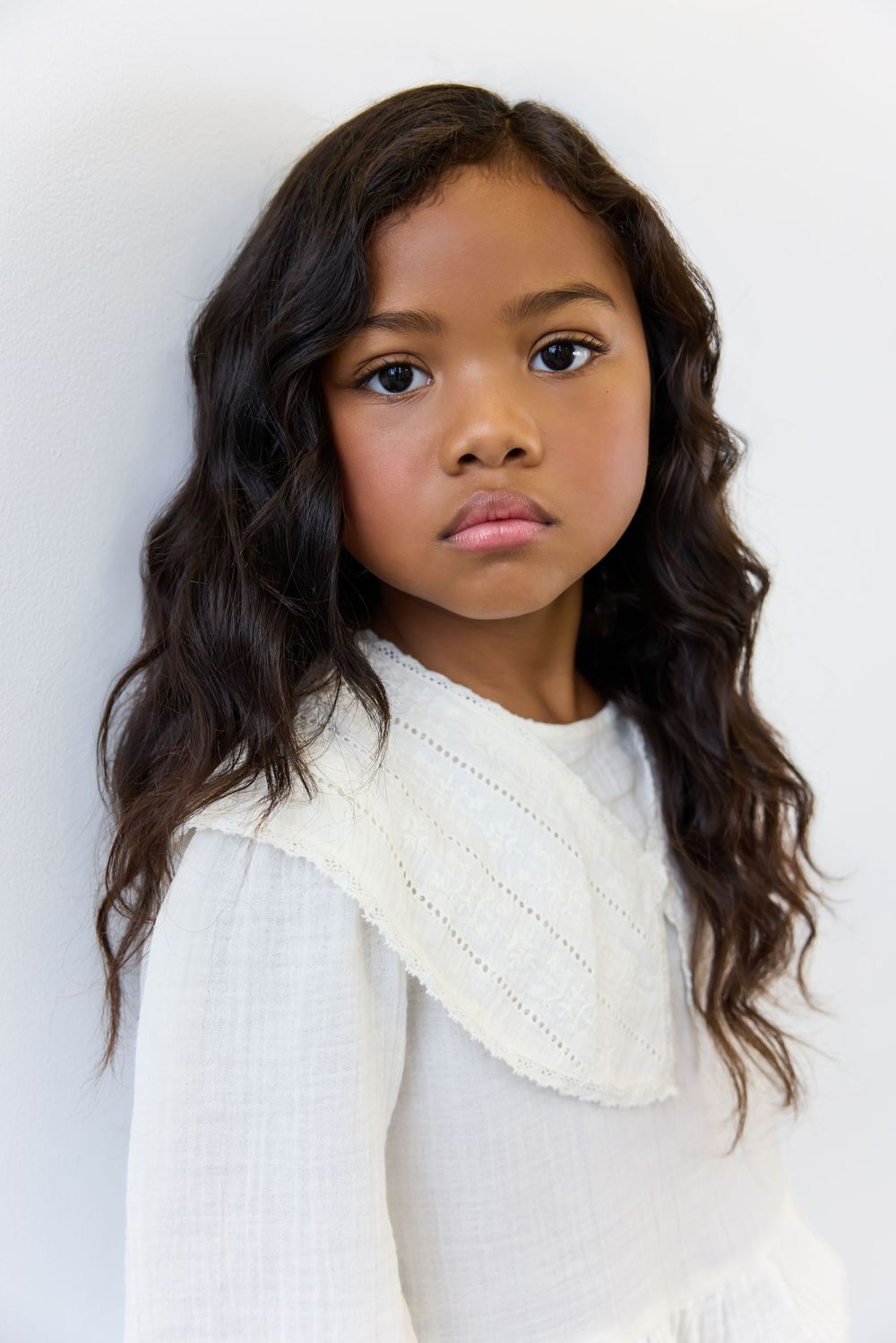 Young girl with long wavy dark hair wearing a white knit outfit poses against a light background.