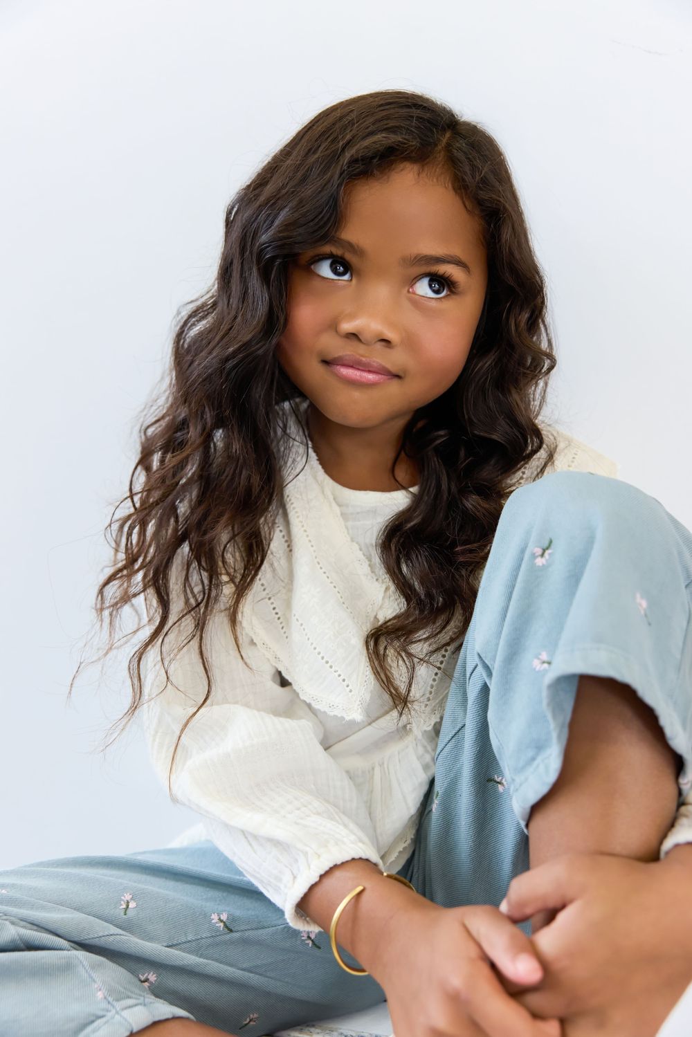 Young girl with long curly hair sitting cross-legged, wearing white top and light blue pants, looking up thoughtfully.