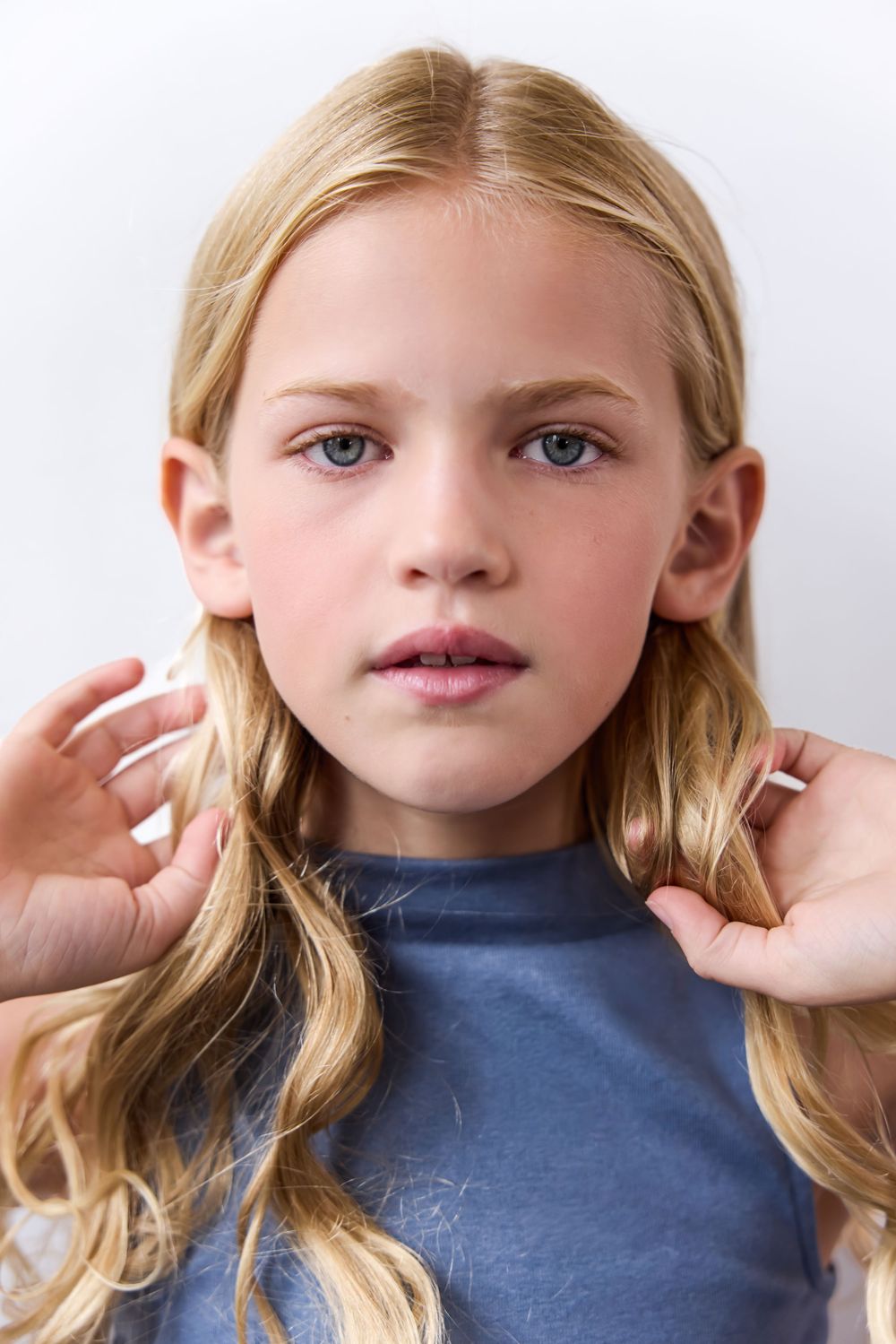 Young blonde girl with blue eyes holding her hair, wearing a blue top, against a white background.