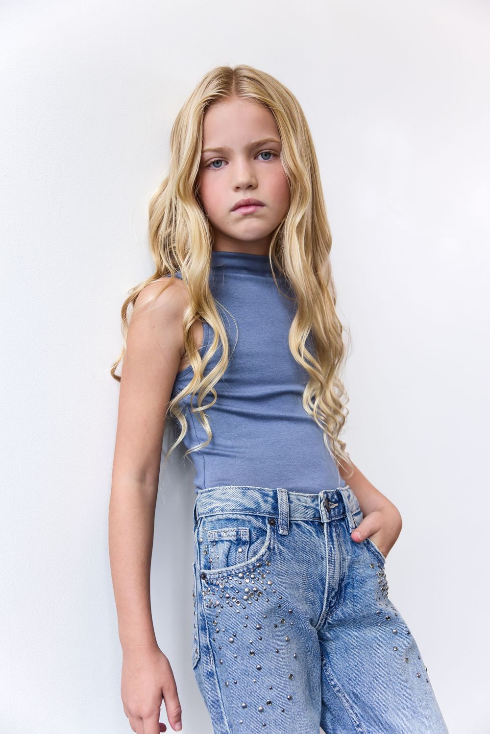 Young girl with long blonde wavy hair wearing a blue sleeveless top and embellished jeans posing against white background.
