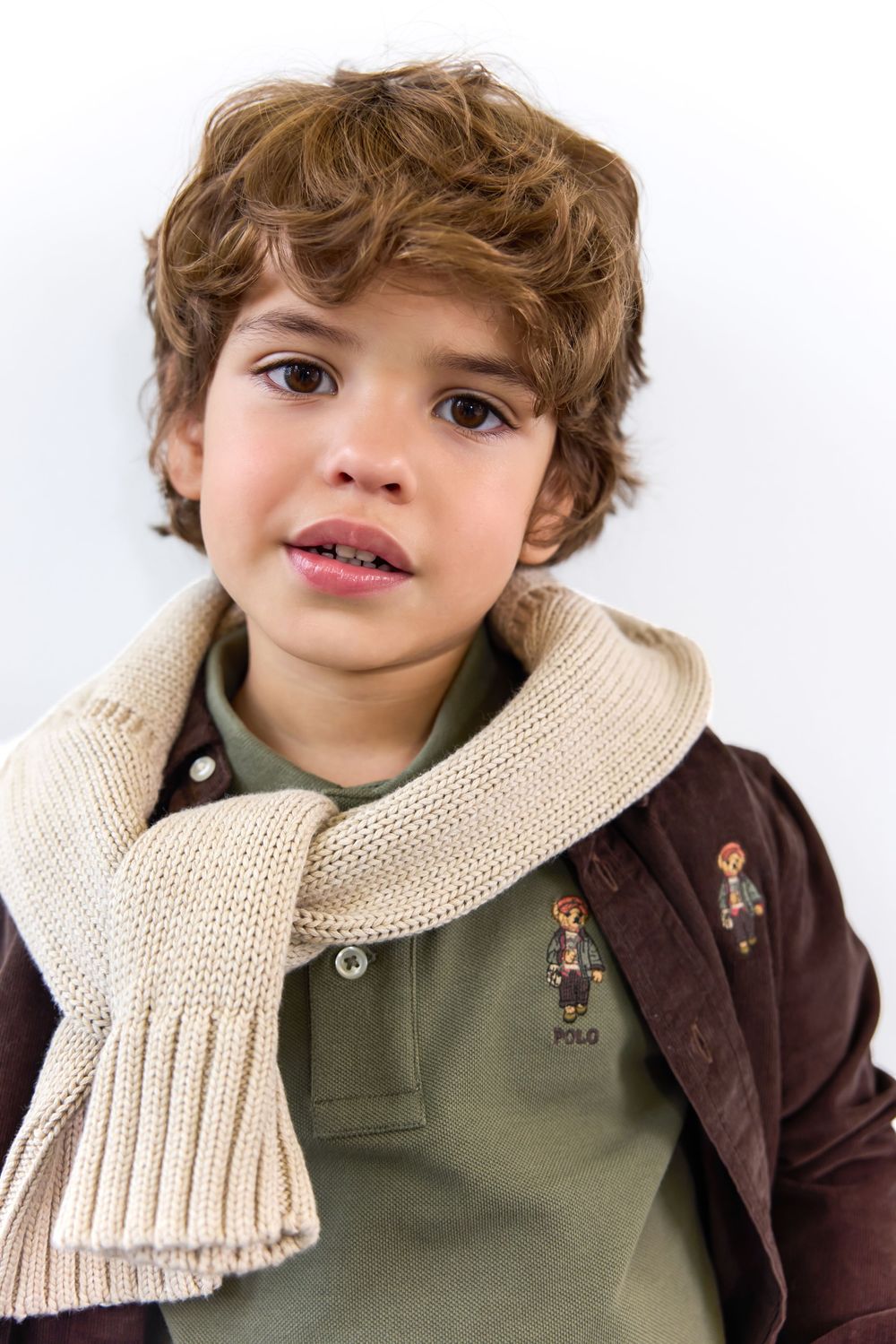 Young boy wearing a green Polo Ralph Lauren shirt, brown jacket, and cream knit scarf against white background.