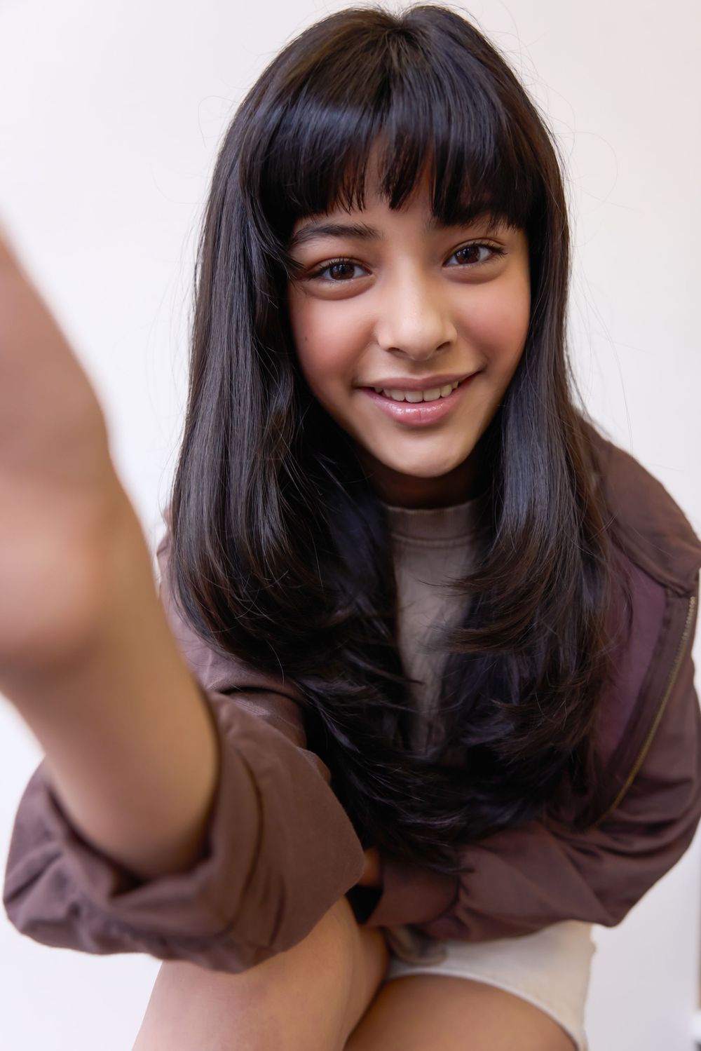 Smiling young girl with long black hair and bangs taking a selfie wearing a brown oversized jacket.