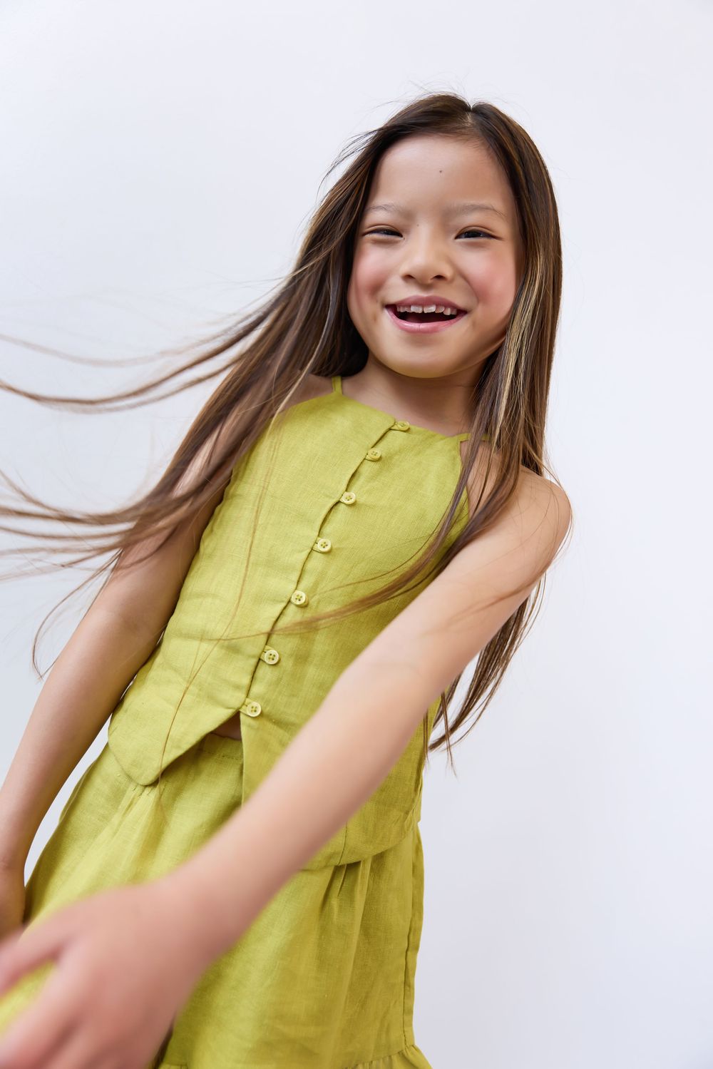 Cheerful Asian girl with long hair twirling in olive green linen dress against white background.