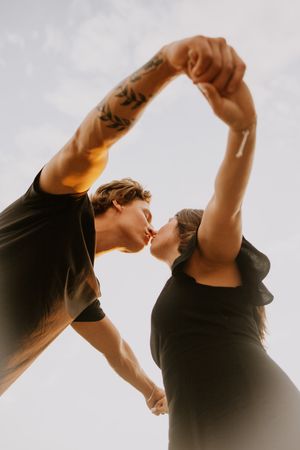Couple in black outfits kissing while holding hands raised overhead, man with leaf tattoo, shot from below against sky.