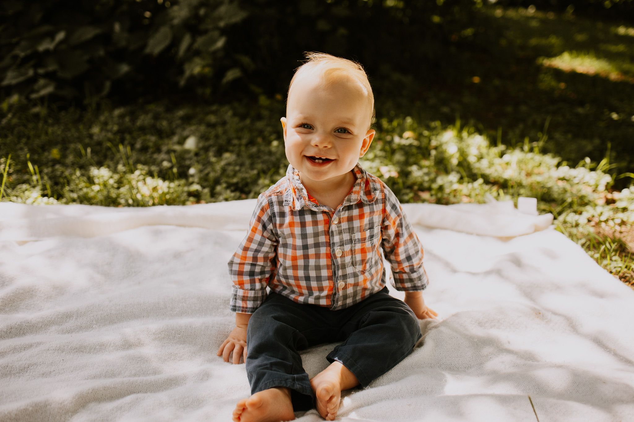 Smiling baby boy sitting on white blanket outdoors in plaid shirt and dark pants, enjoying sunny day in green garden.