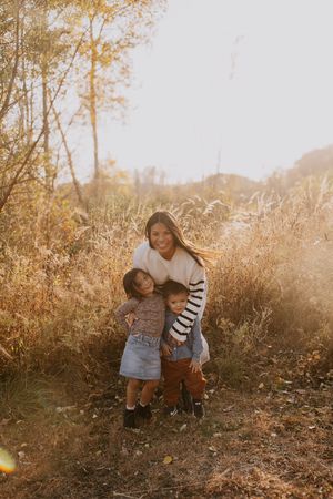 A mother smiles warmly while hugging her two young children in a sunlit autumn field with golden foliage.