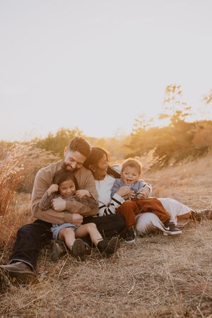 Family of four sitting together in golden field at sunset, parents hugging children, warm autumn light.