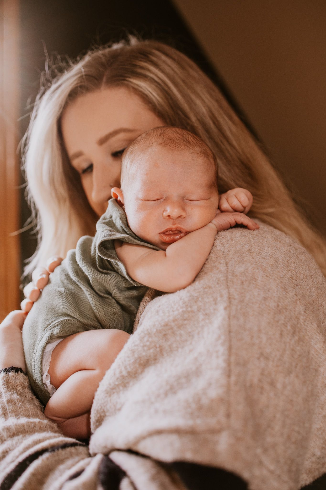 A mother with blonde hair gently holds her sleeping newborn baby dressed in a green onesie against her shoulder.