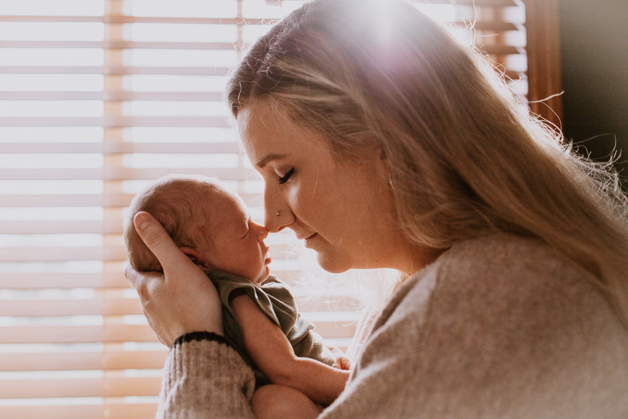 A mother tenderly touches foreheads with her newborn baby, bathed in warm sunlight near window blinds.