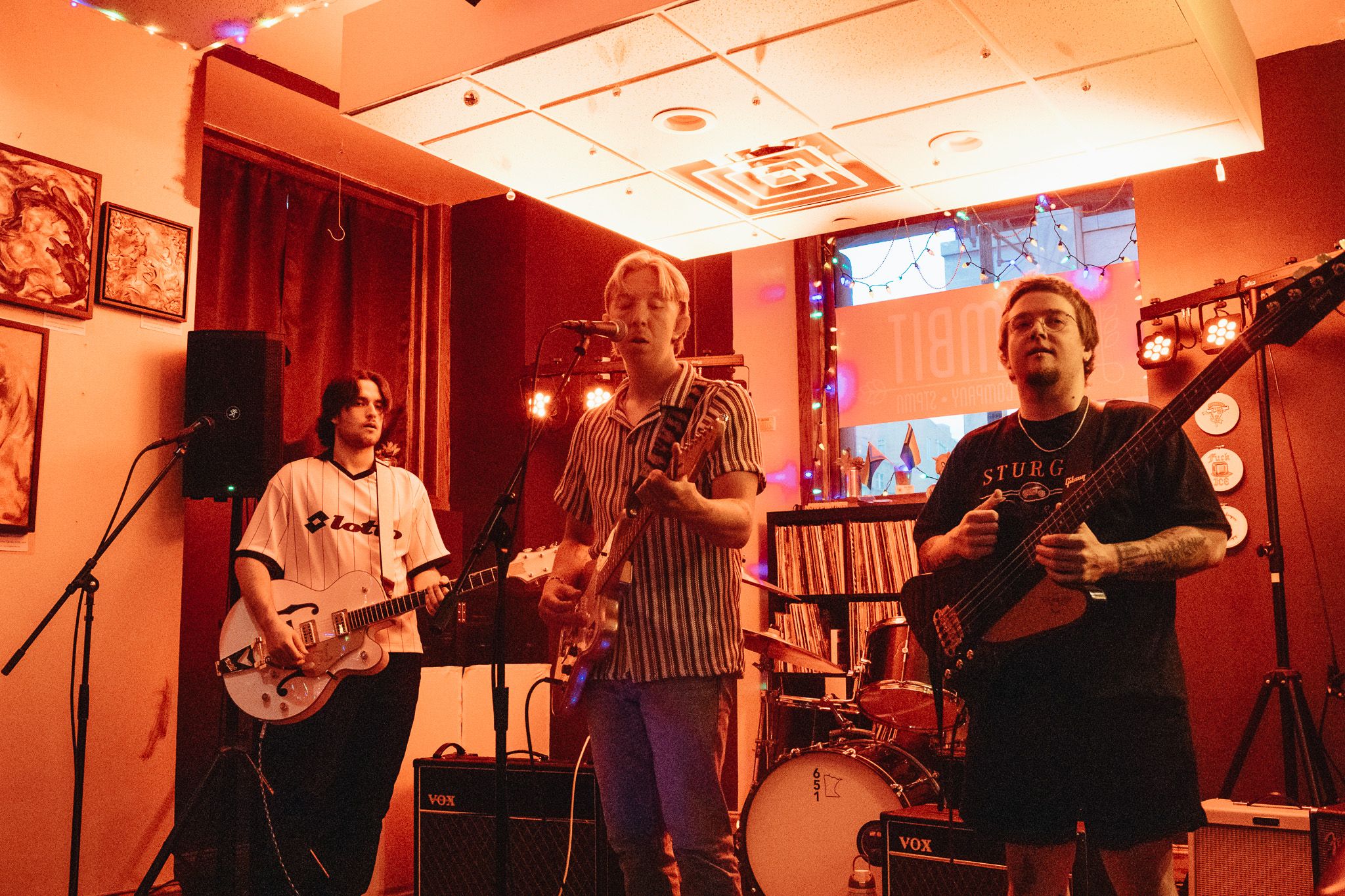 Three musicians perform in a cozy red-lit venue, playing electric guitars with a drum set in the background.