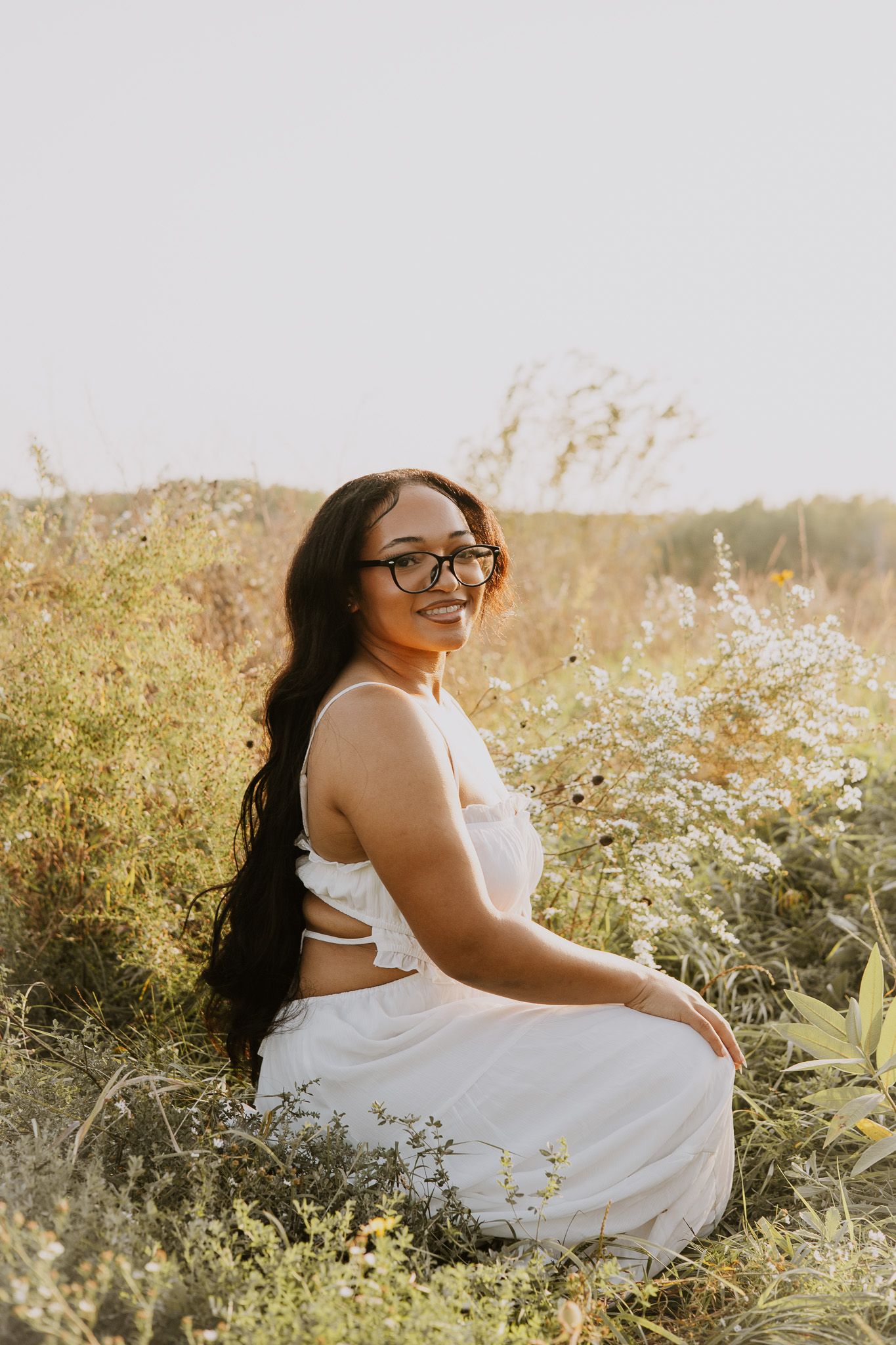 Young woman in white dress sitting in wildflower field, smiling, wearing glasses, long dark hair, golden hour light.