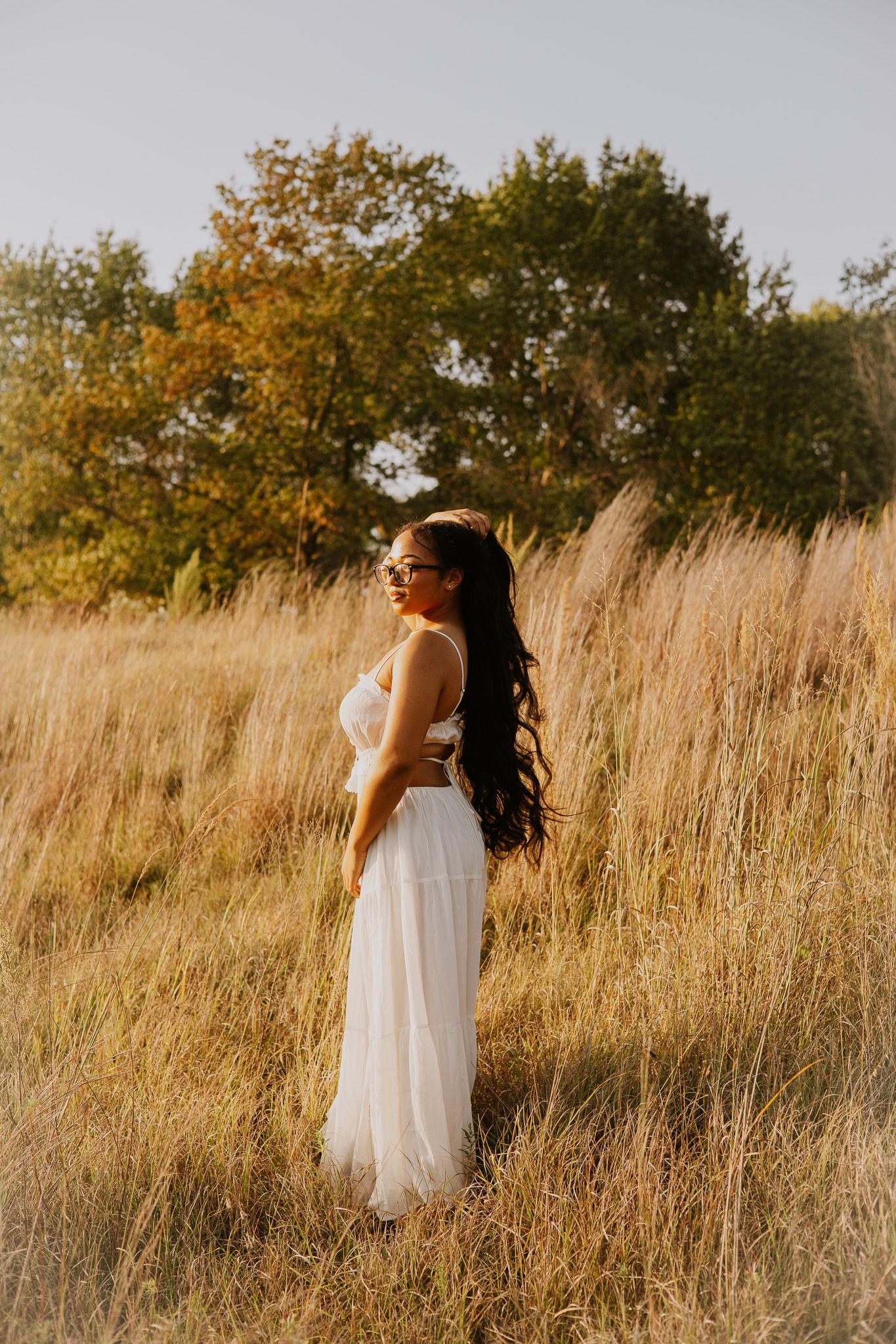 Woman in white dress standing in golden field with trees, long dark hair, sunglasses, outdoor nature photography.