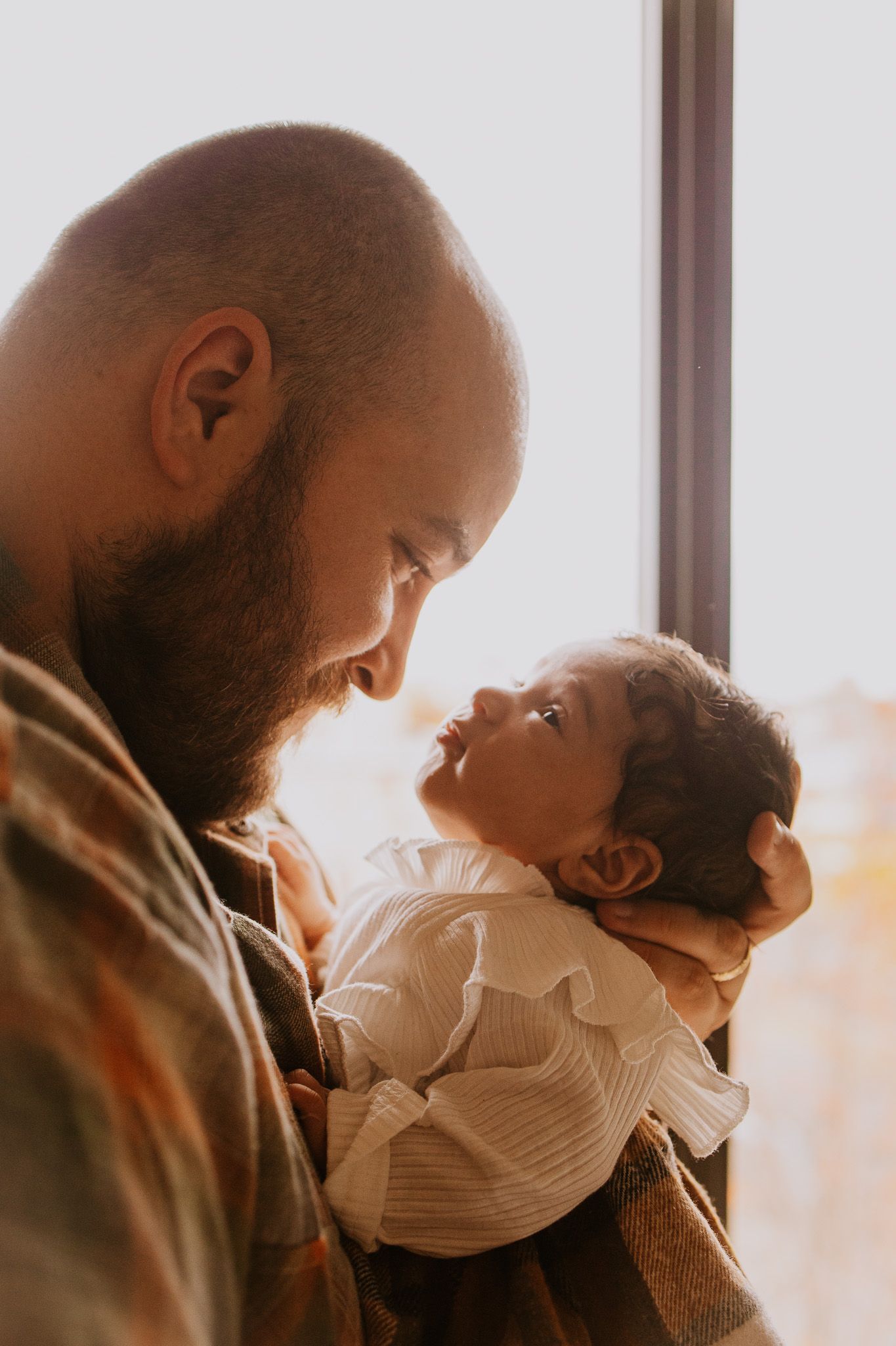 Bearded father tenderly holds his newborn baby near a sunlit window in a warm, intimate portrait.