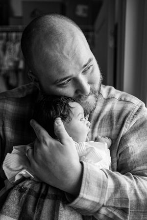 Black and white photo of a bald bearded father tenderly holding and kissing his newborn baby.