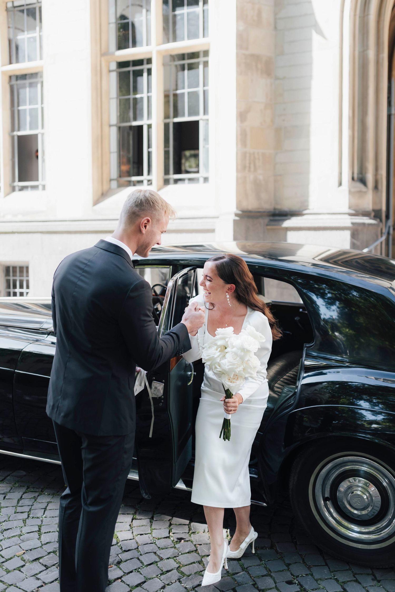 Elegant bride in white midi dress holding bouquet steps out of black luxury car assisted by groom in dark suit.
