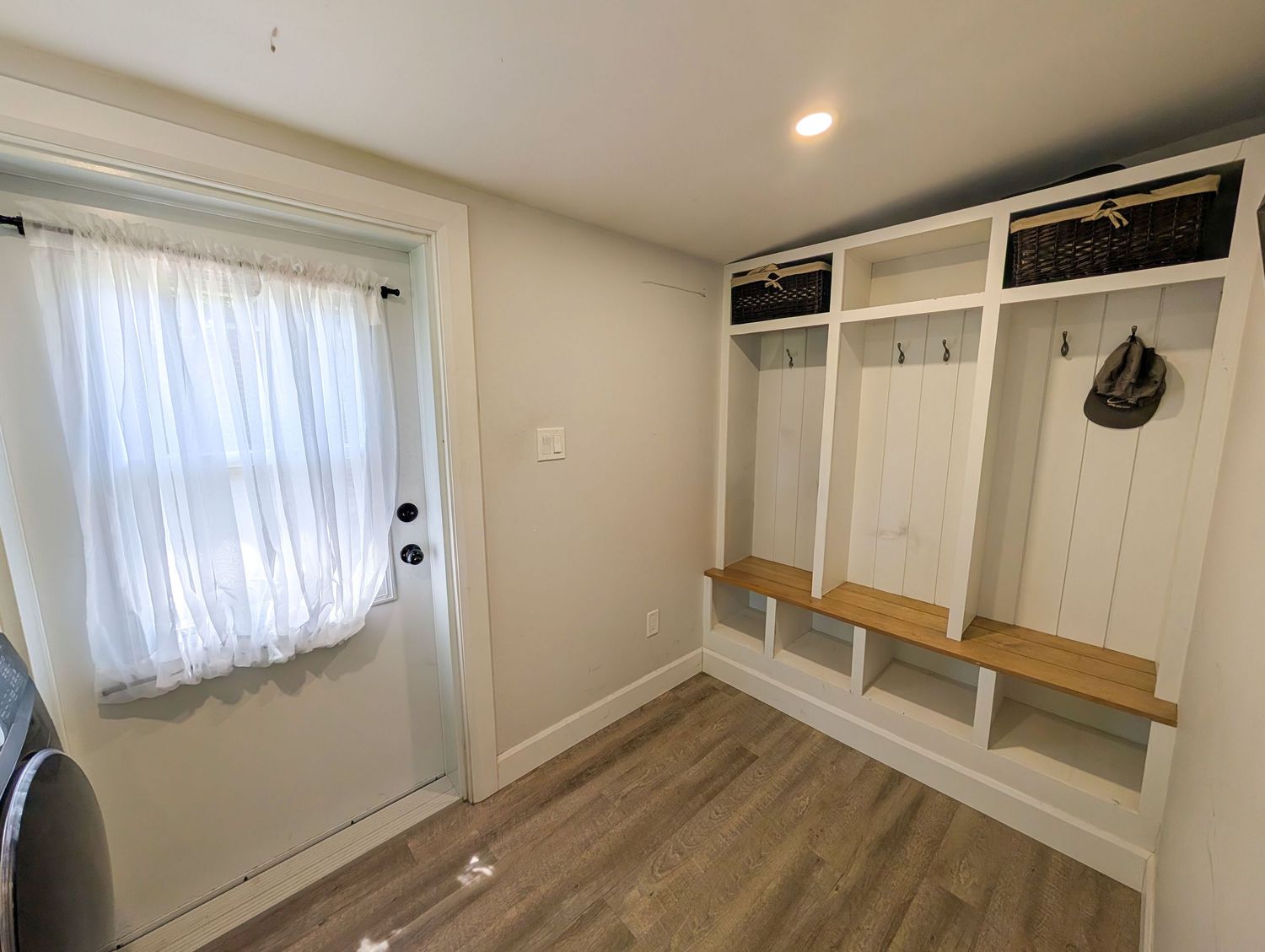 Bright mudroom with white built-in lockers, wooden bench, coat hooks, and shiplap panels beside a white door with curtain.