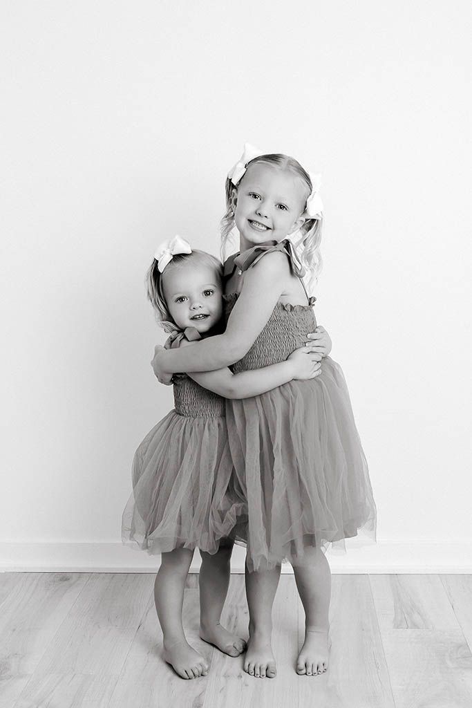 Two young sisters in matching tulle dresses and hair bows share a sweet hug in a black and white portrait.