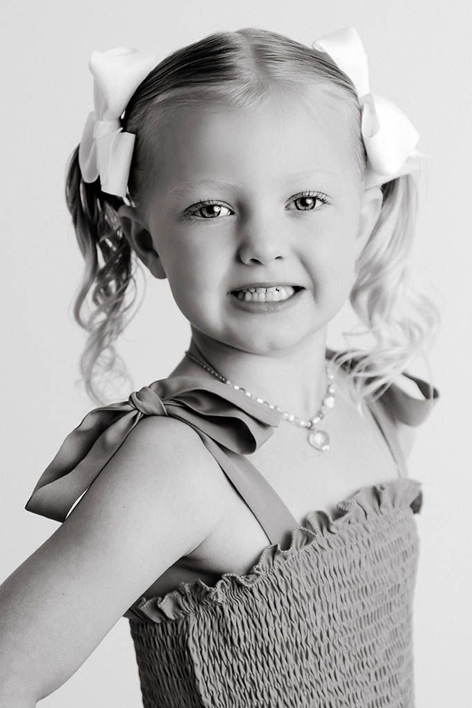 Black and white portrait of a smiling young girl with pigtail bows wearing a ruffled dress and pearl necklace.