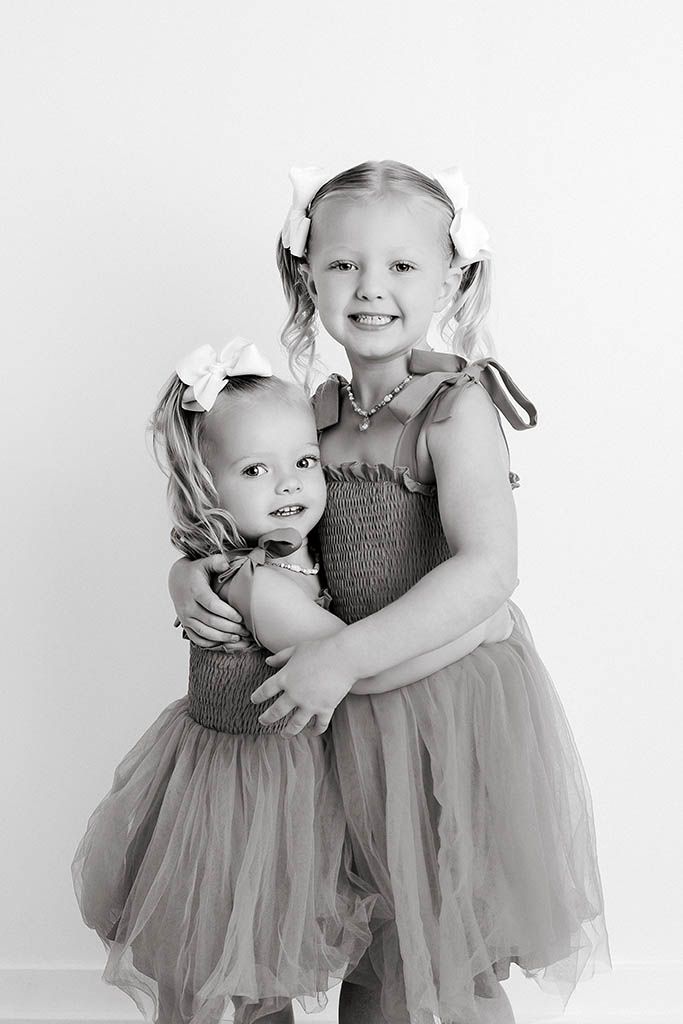 Two sisters in matching smocked dresses with bow hair accessories posing together in black and white photo.