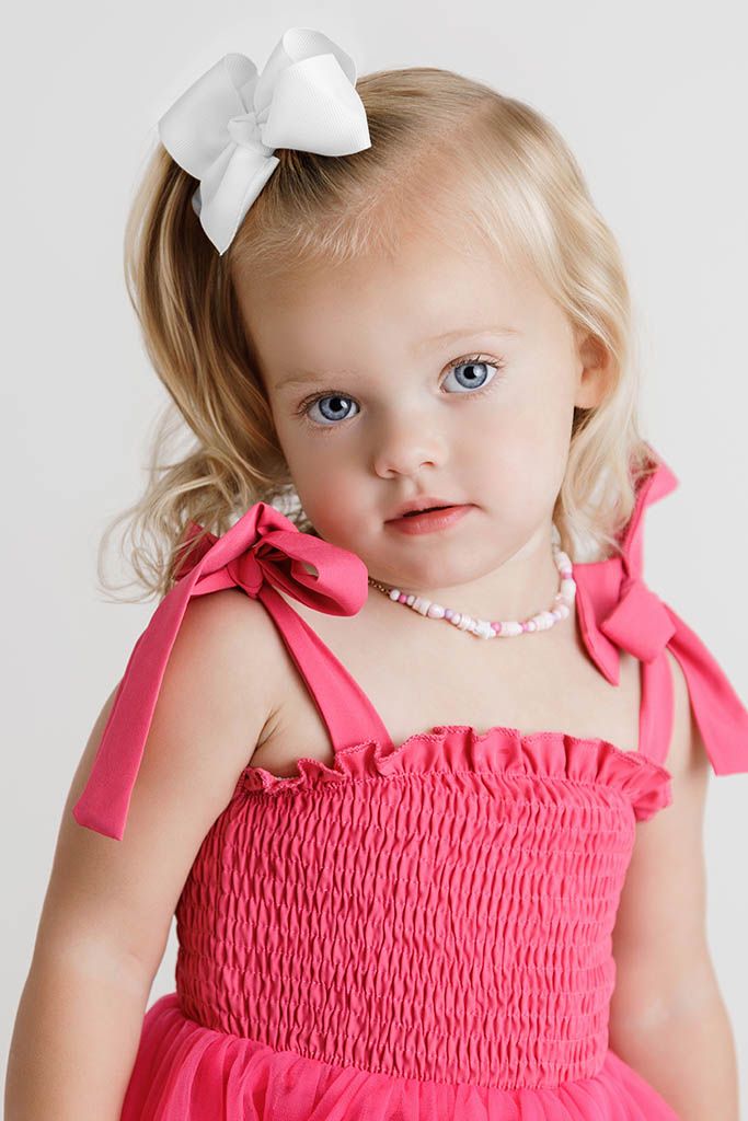 Toddler girl with blue eyes in pink smocked dress with bow hair accessory and beaded necklace.