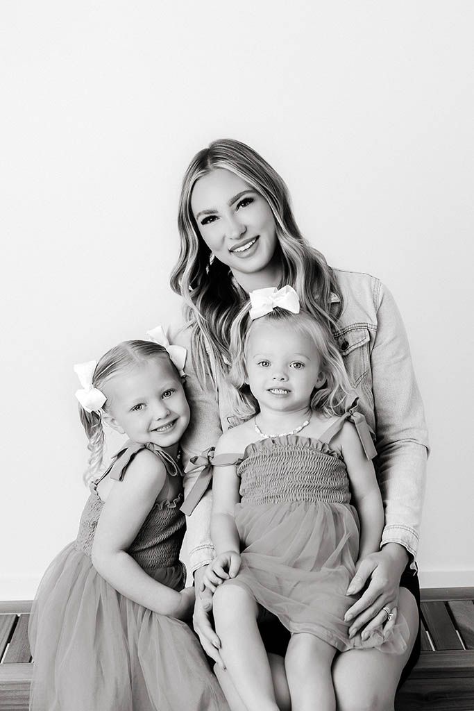 Black and white portrait of a smiling mother with two young daughters wearing bows, seated together on a bench.