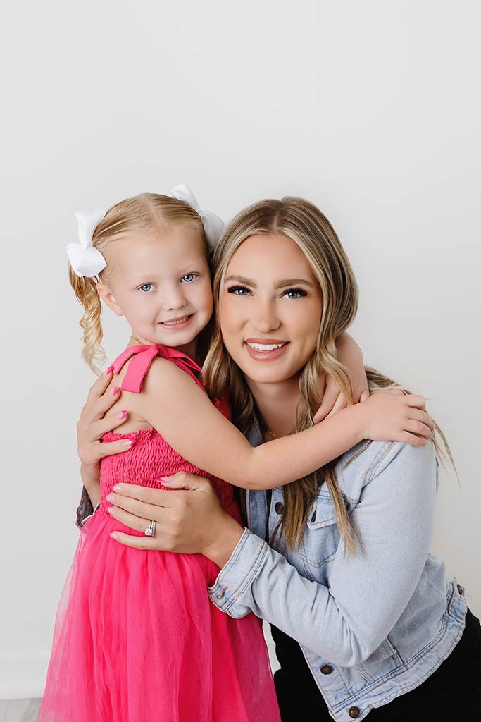 A smiling woman in a denim jacket hugs a young girl in a pink tutu dress with a white bow in her blonde hair.