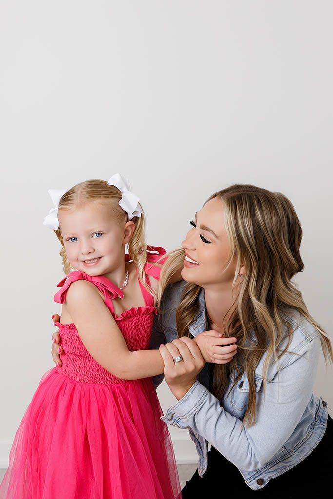 A woman in a denim jacket smiles at a young girl in a pink tutu dress with a white bow, posing together.