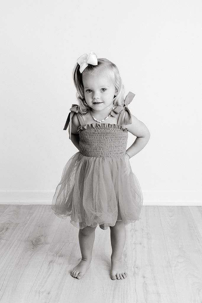 Black and white photo of a cute toddler girl in a tutu dress with bow accessories, standing barefoot in a studio.