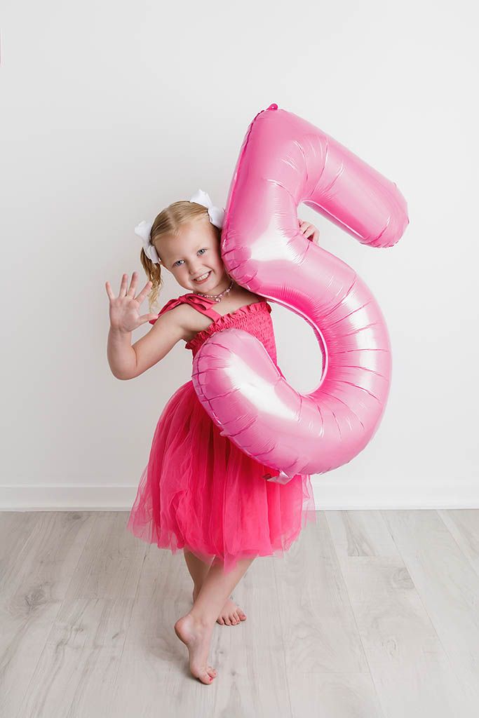 A cheerful young girl in a pink tutu dress holds a large pink number 5 balloon, waving at the camera.