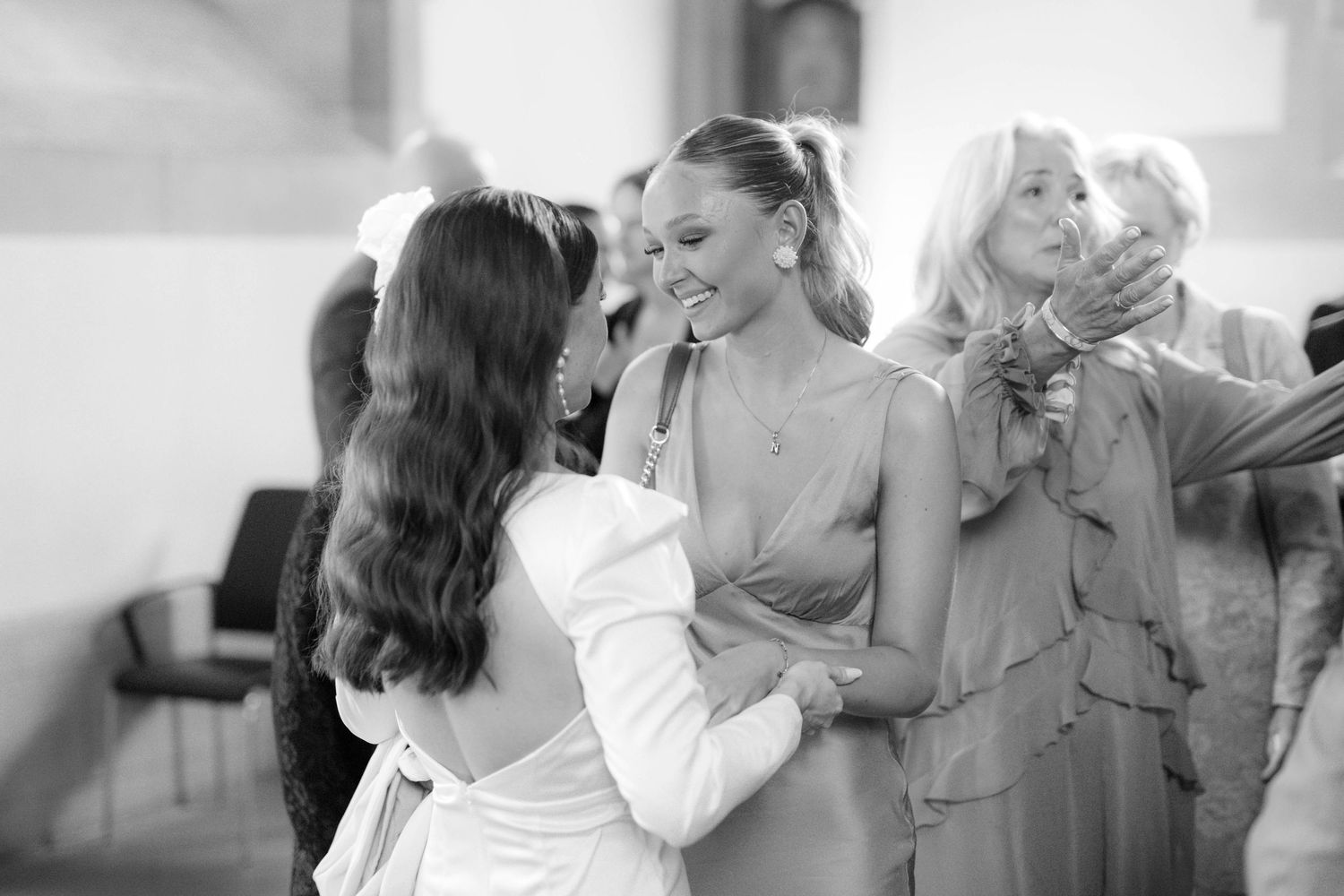 Black and white photo of elegantly dressed women smiling and embracing at a formal event.