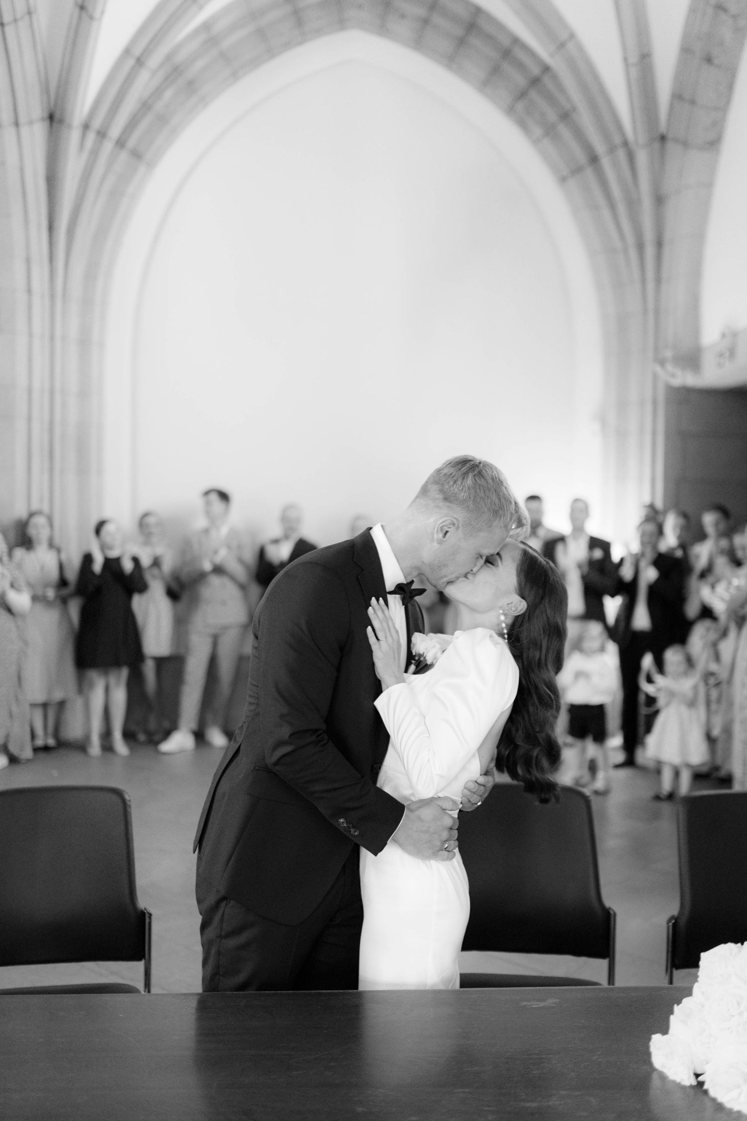 Black and white photo of a couple sharing their first kiss at a wedding ceremony in an elegant arched venue.