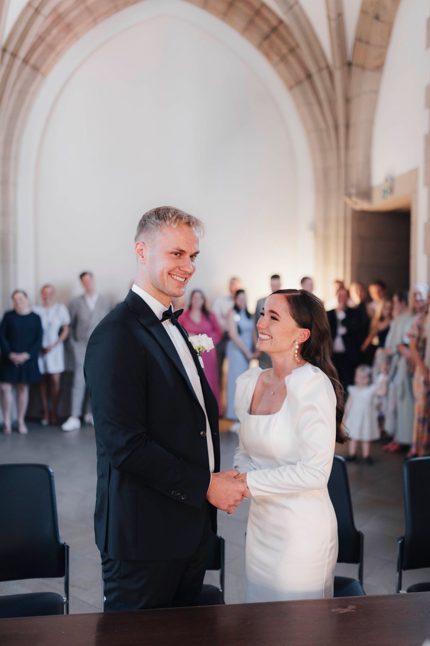 A smiling bride and groom hold hands at their wedding ceremony in an elegant vaulted hall with guests in background.