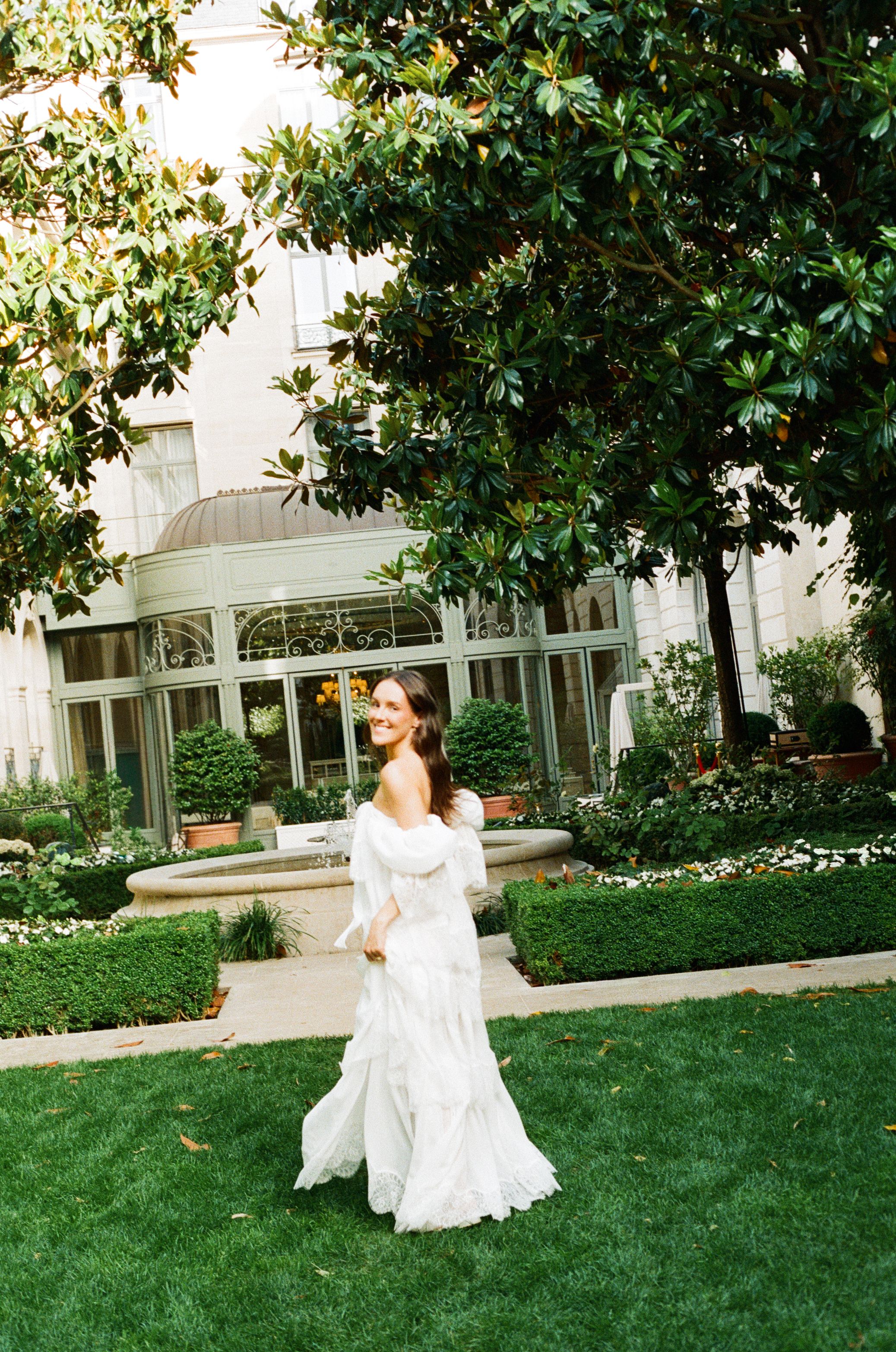 Bride in white off-shoulder gown twirling in lush garden courtyard outside elegant building.