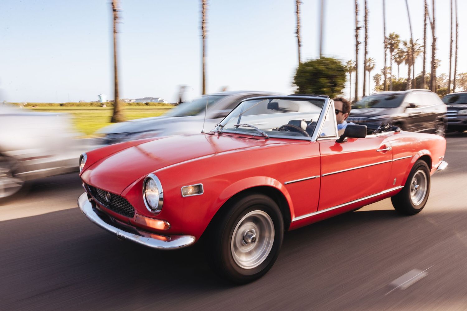 Red vintage Fiat 124 Spider convertible cruising along a palm-lined coastal road on a sunny day.