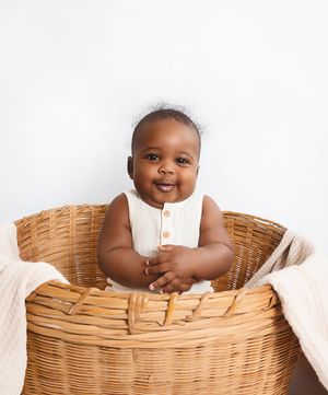 Smiling baby sitting in a large wicker basket wearing a cream romper against a white background.