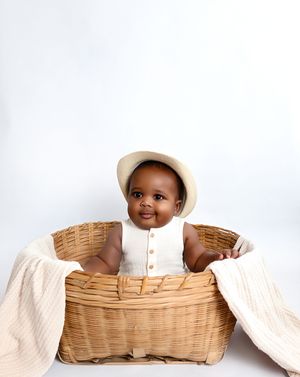 Smiling baby in a cream outfit and hat sitting in a wicker basket with soft white fabric.