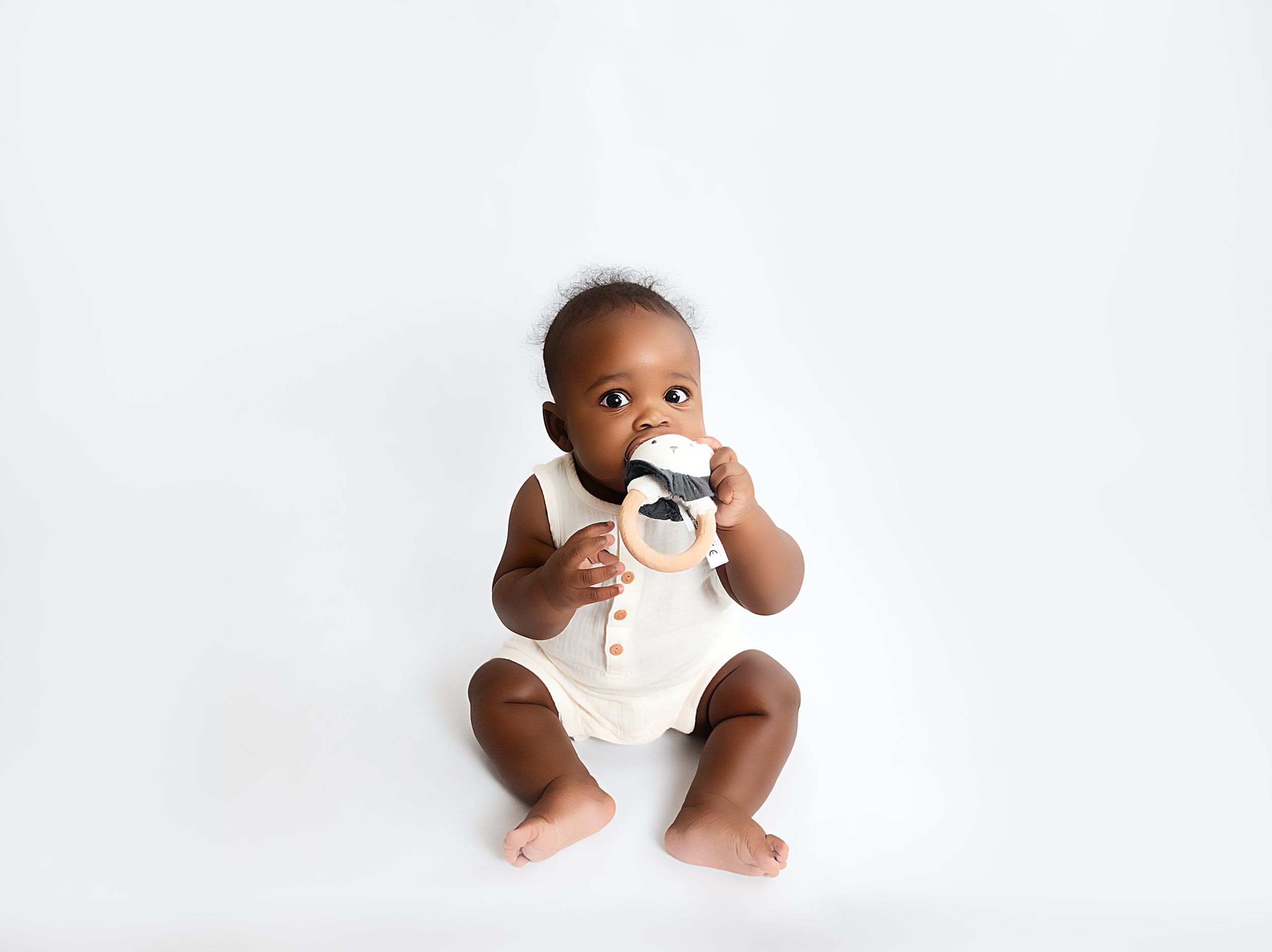 A cute Black baby in a cream romper sits on a white background, chewing on a black and white teething toy.