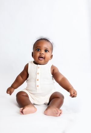 Adorable baby in cream sleeveless romper with wooden buttons sits on white background, looking up curiously.